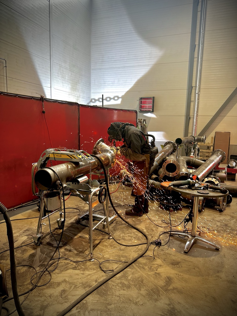 A welder working on a pipe in a fabrication workshop, with multiple prefabricated pipe sections neatly stacked on the right, ready for further processing and assembly.